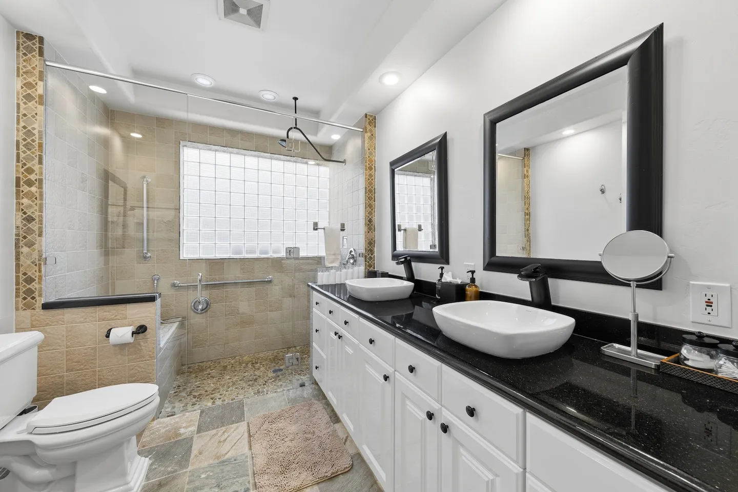 Bathroom with a double-sink vanity with black countertop, toilet, and walk-in shower with glass block window.
