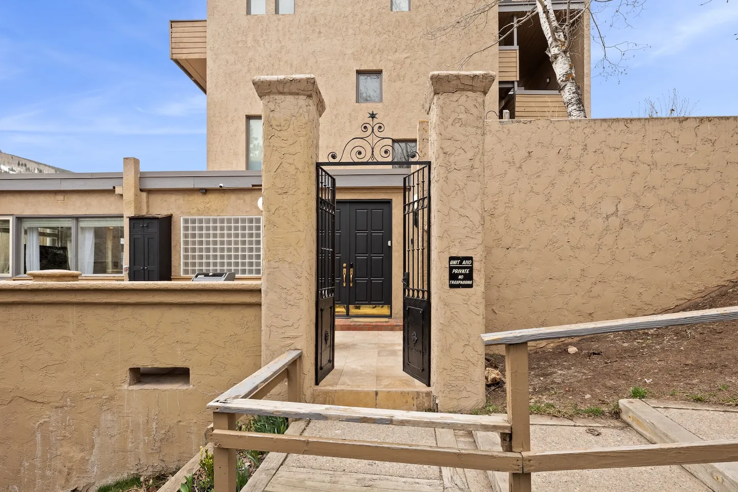 Exterior gated entrance with open wrought iron gates leading down steps toward a black double front door.