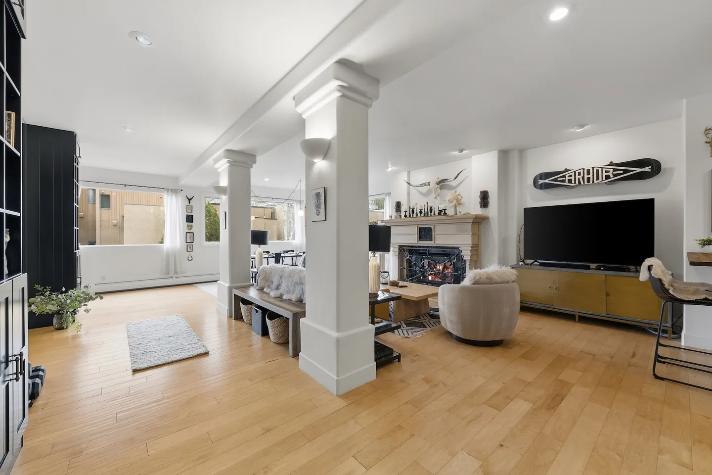 Open-concept living room with light wood floors, support columns, a fireplace, and a television above a media console.