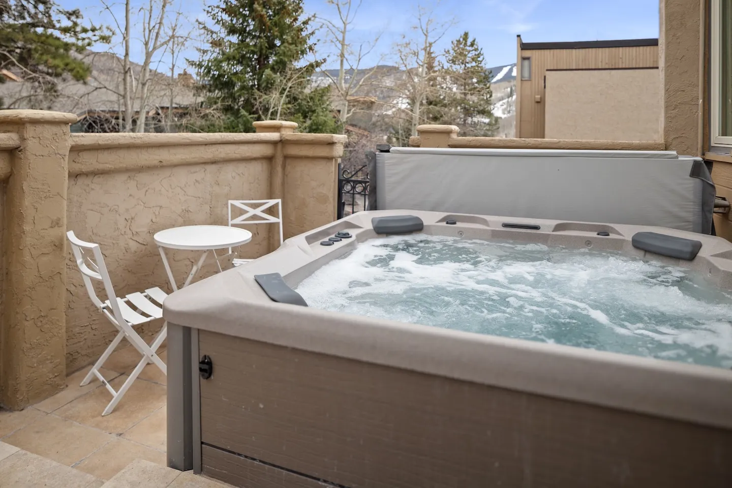 Outdoor hot tub bubbling on a stone patio with a small table and chairs nearby and trees in the background.