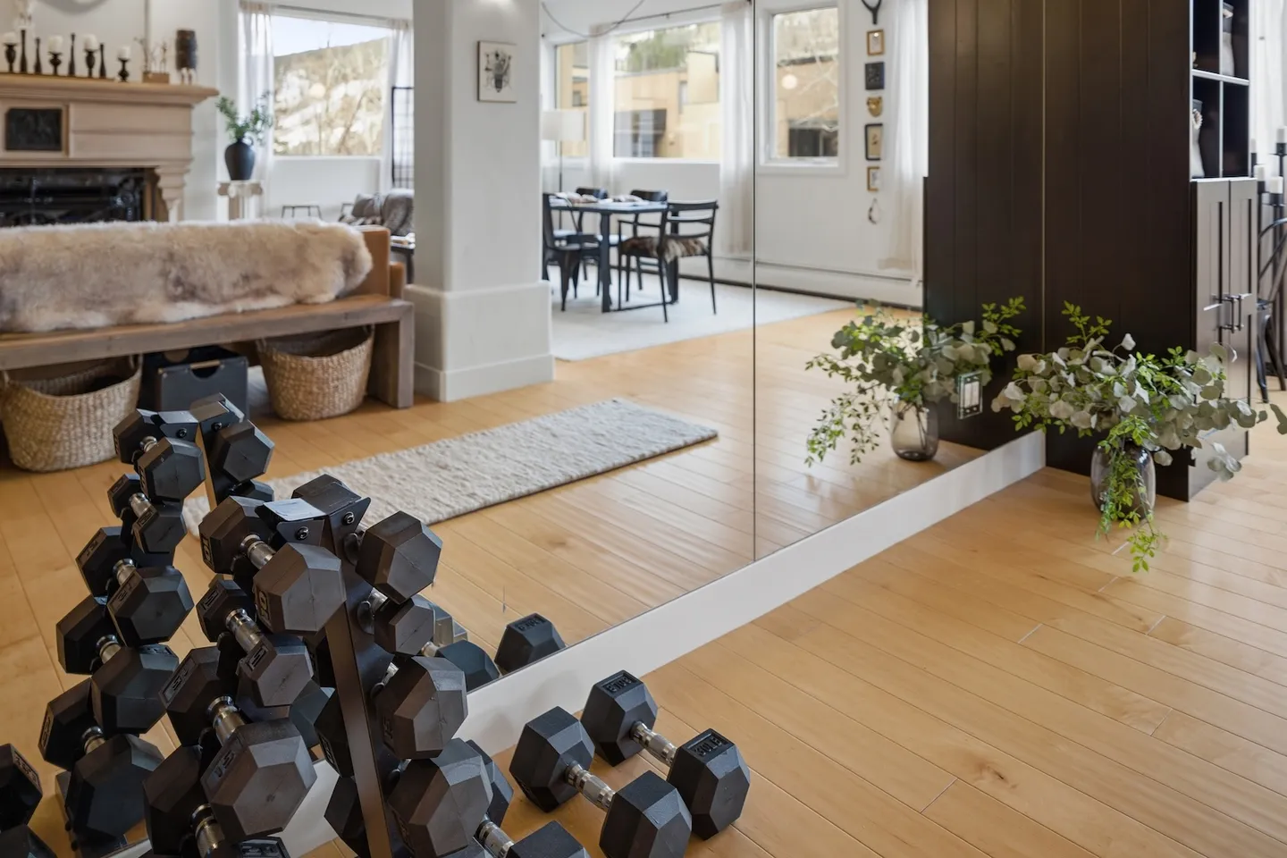 A rack of black dumbbells sits on a light wood floor in front of a large mirror reflecting an open-concept living and dining area.