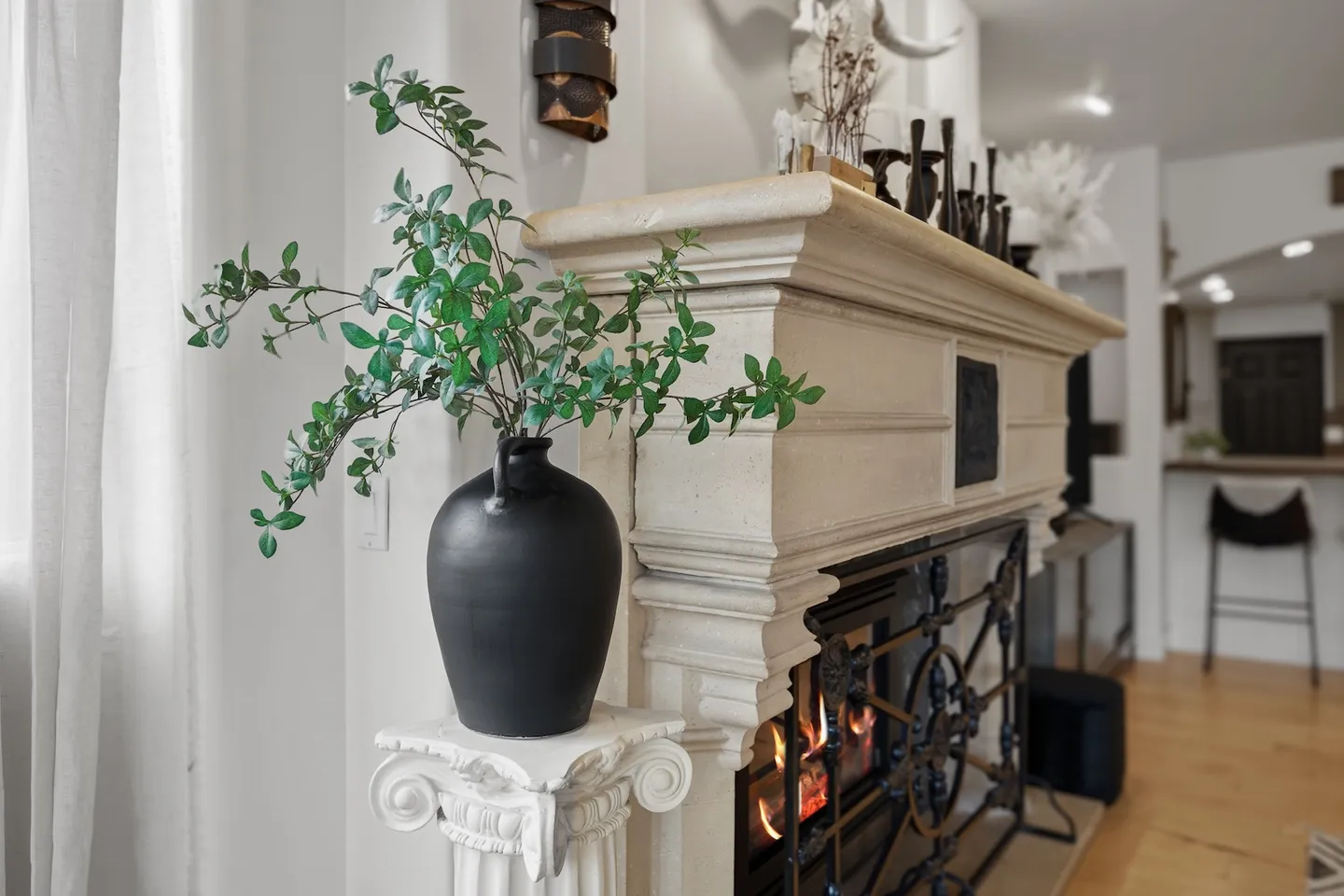 Close-up of a lit fireplace with an ornate stone mantel, featuring a black vase with greenery on a decorative pedestal in the foreground.