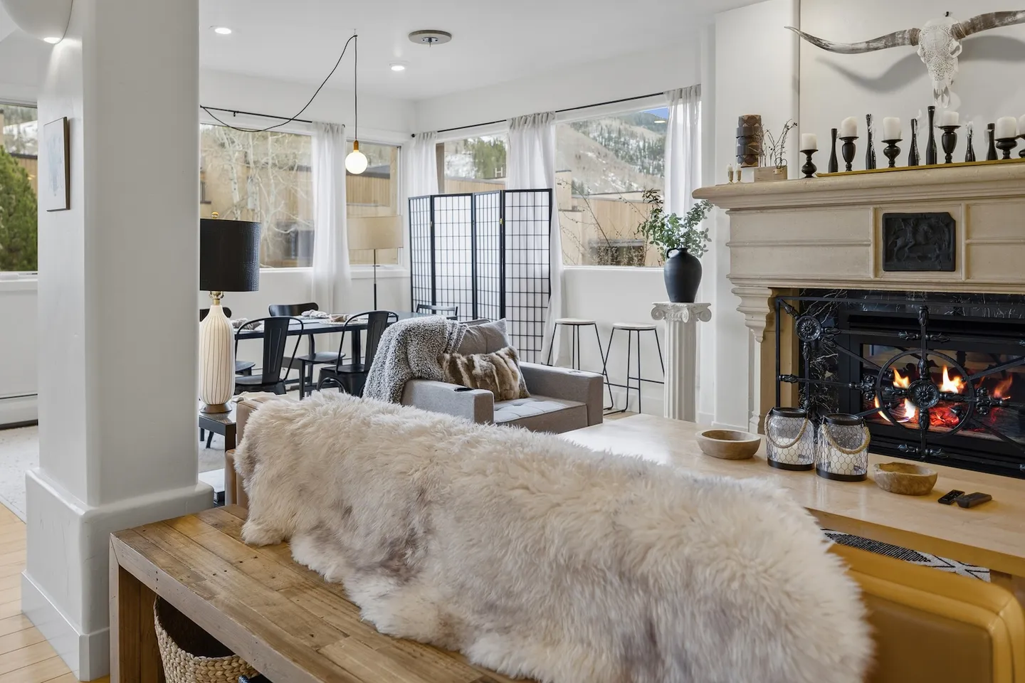 Open-concept living room featuring an ornate stone fireplace with a fire burning, large windows with mountain views, and adjacent dining area.
