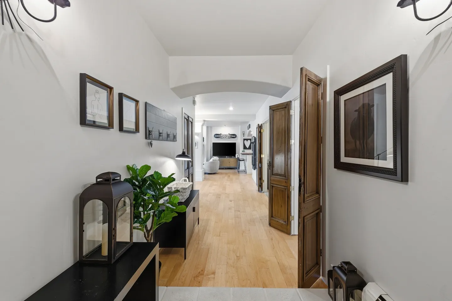 A long interior hallway with wood floors leading toward a living area, decorated with wall art, a console table with a lantern, and wooden panel doors.