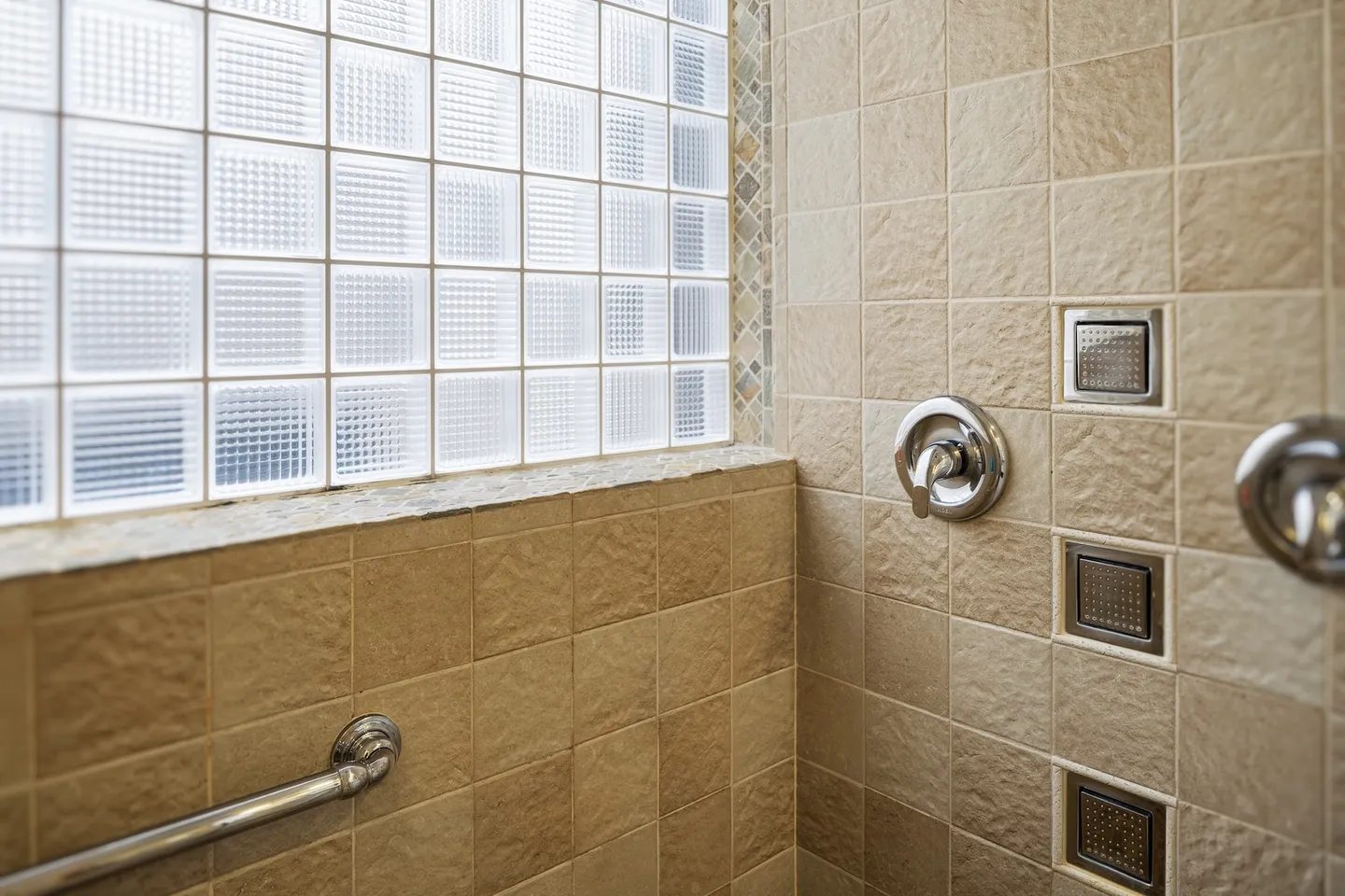 Tiled walk-in shower corner featuring glass block window, chrome fixtures, and body spray jets.
