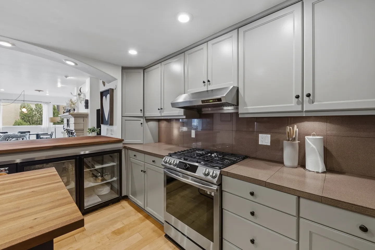 Kitchen with gray cabinetry, brown tiled countertops and backsplash, stainless steel gas range and hood, and under-counter beverage cooler.