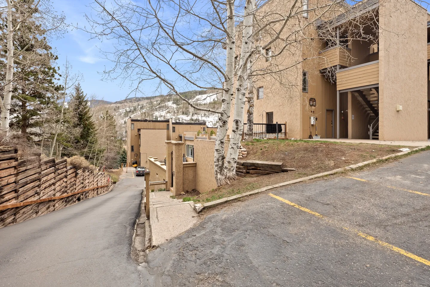 Exterior view of multi-story tan stucco condominium buildings along a sloped paved driveway with mountains in the background.