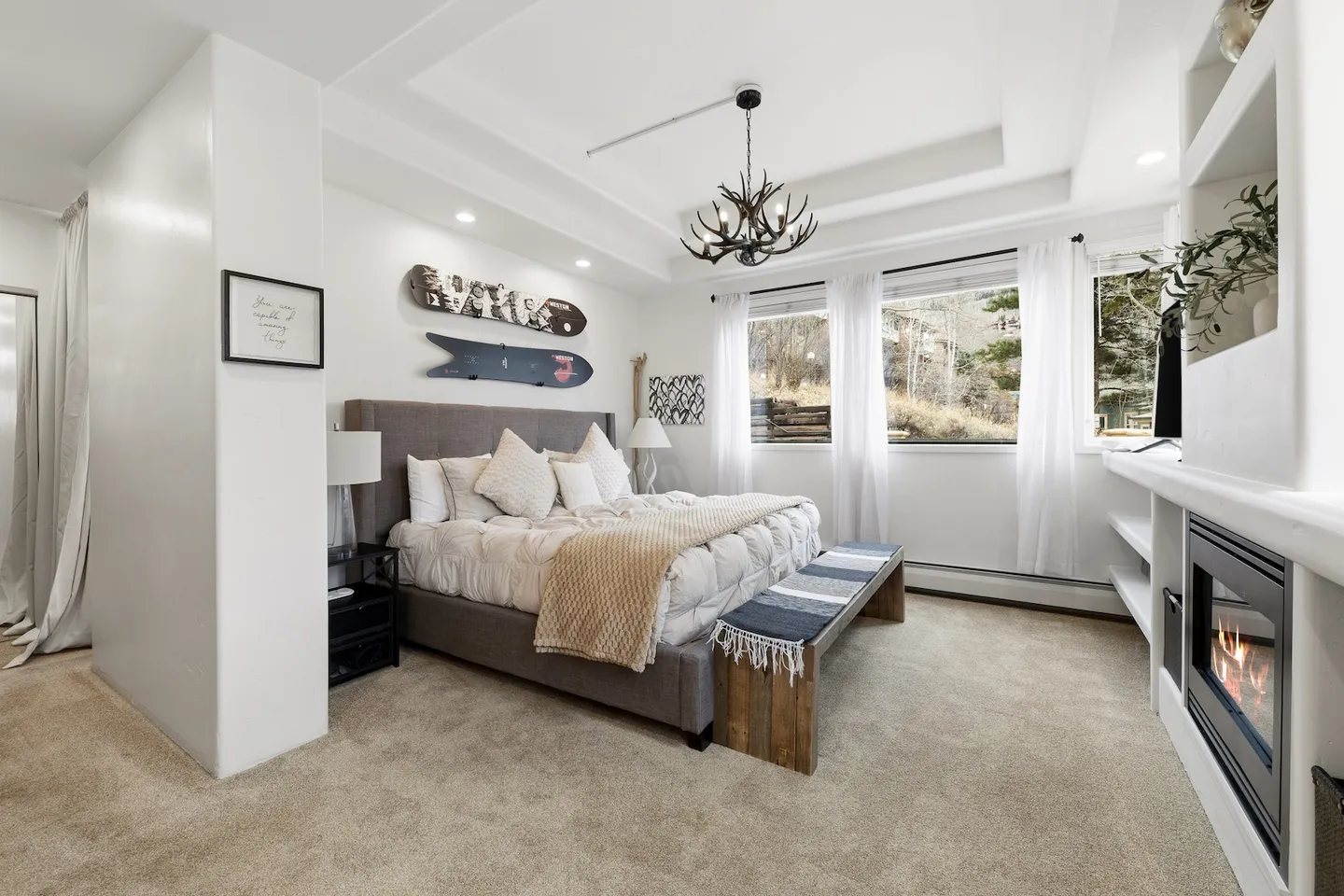 Bedroom with an upholstered bed, antler chandelier, carpeted flooring, and a built-in fireplace with a television above.