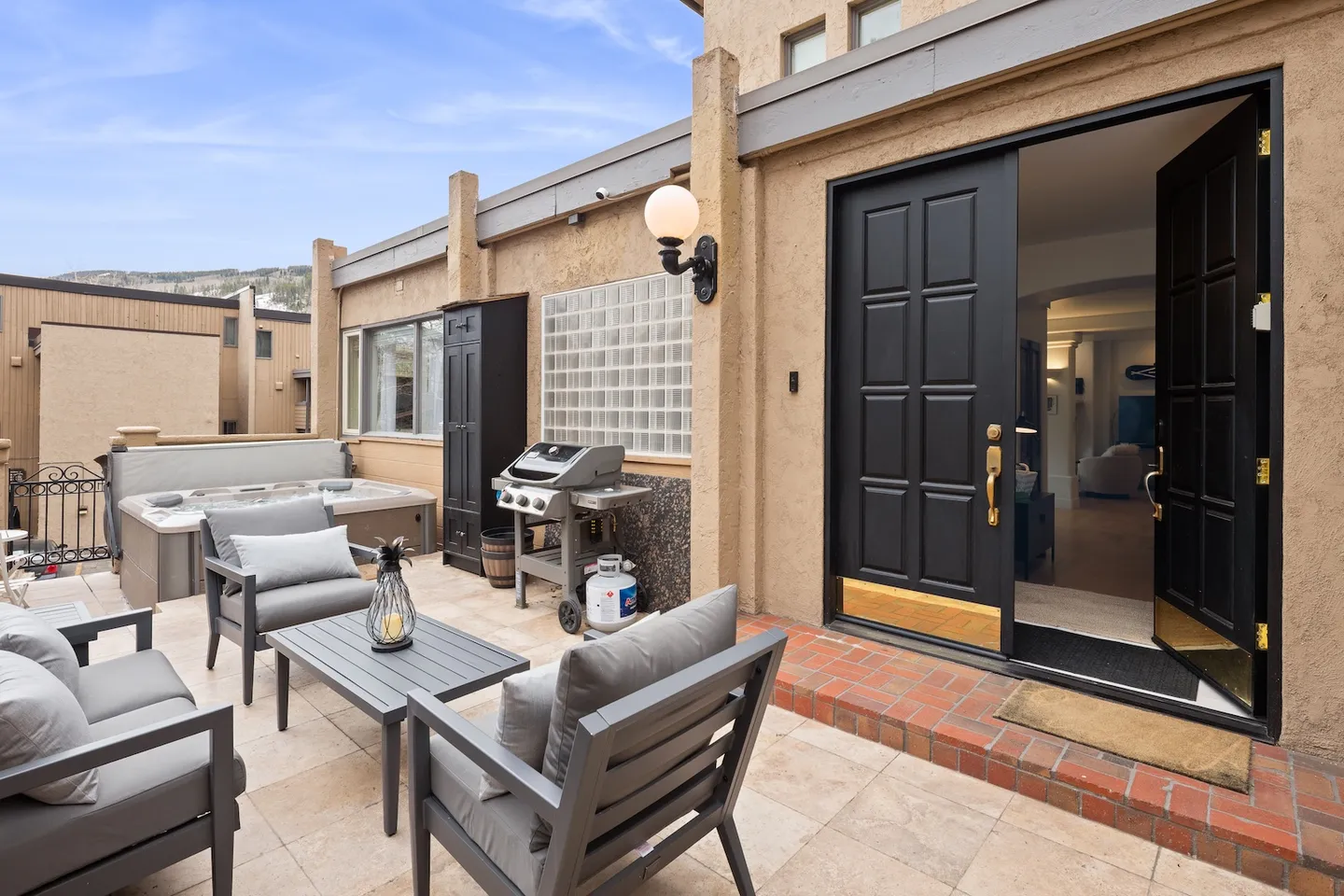Outdoor tiled patio with outdoor lounge seating, a barbecue grill, and a hot tub near an open black double front door.