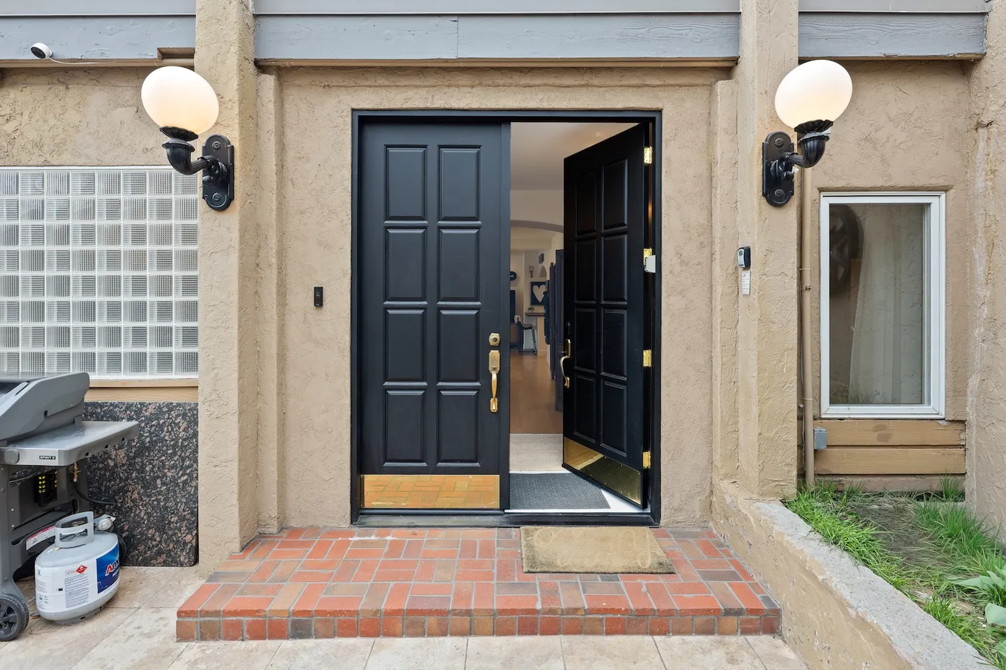Front entrance of a home featuring black paneled double doors with one open, flanked by globe sconces and brick steps.