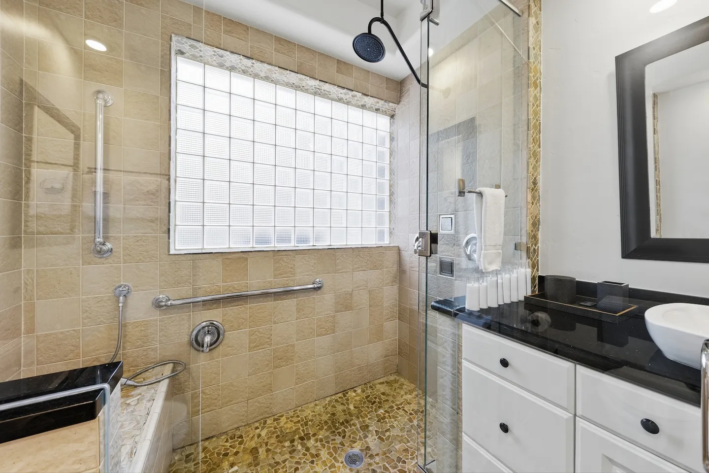 Bathroom with a walk-in glass shower featuring tan tile, a glass block window, pebble floor, and an adjacent vanity with a black countertop and vessel sink.