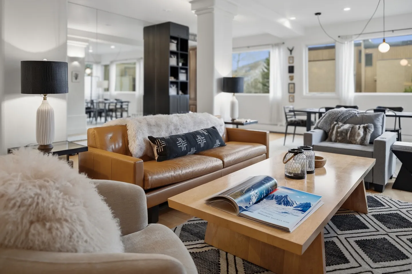 Bright modern living room with a tan leather sofa, wooden coffee table, armchair, and an open dining area in the background.