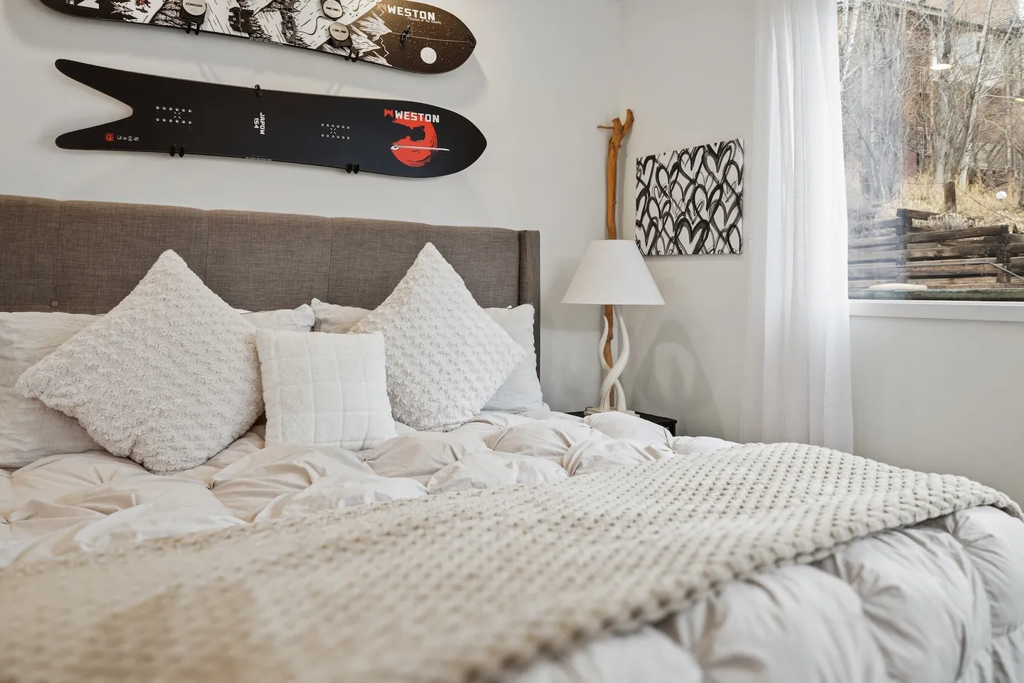 Bedroom with a plush bed, grey fabric headboard, two snowboards mounted on the wall, and a window with sheer curtains.