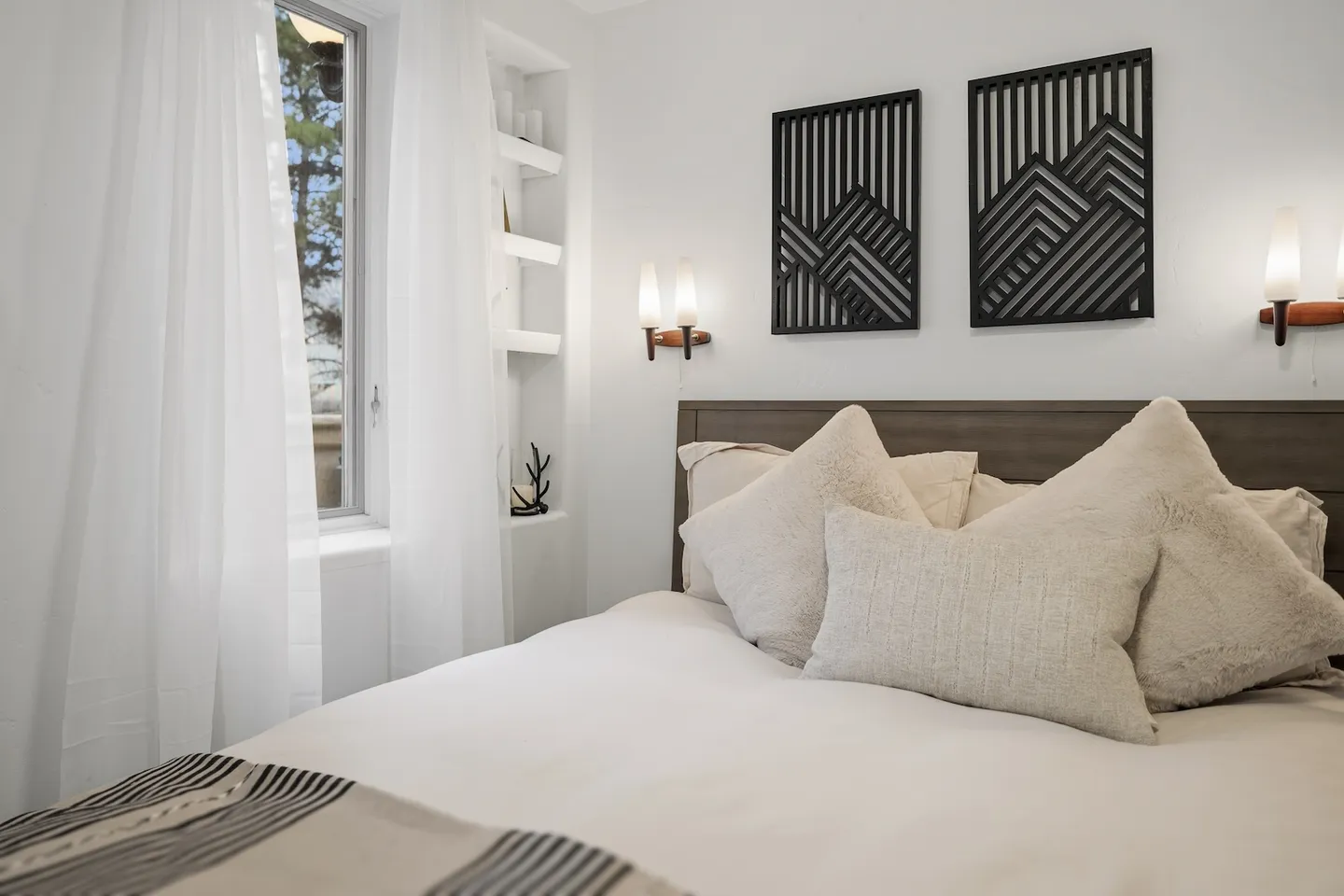 Bedroom with a neatly made bed, accent pillows, wall sconces, geometric art, and a window with sheer curtains.