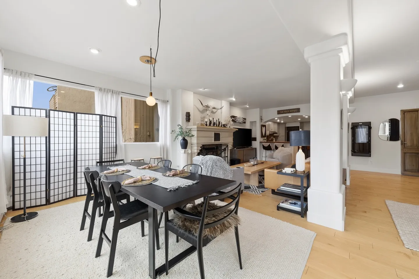 Dining area with a black dining table and chairs on a light area rug, leading into an open living room with a fireplace.
