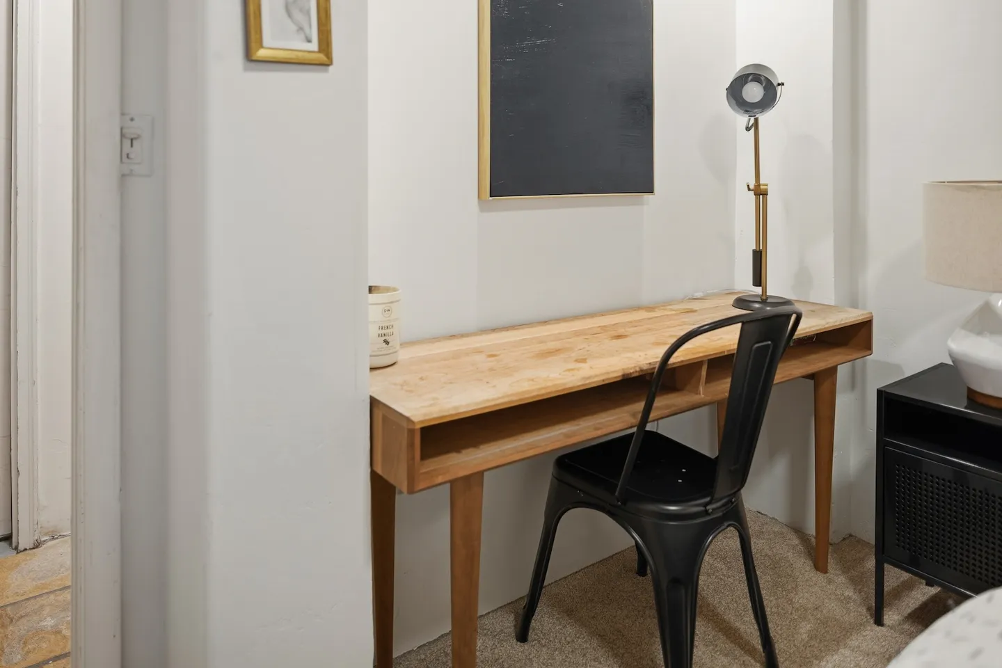A wooden work desk with an open storage cubby, paired with a black metal chair, adjustable desk lamp, and framed artwork on the wall.