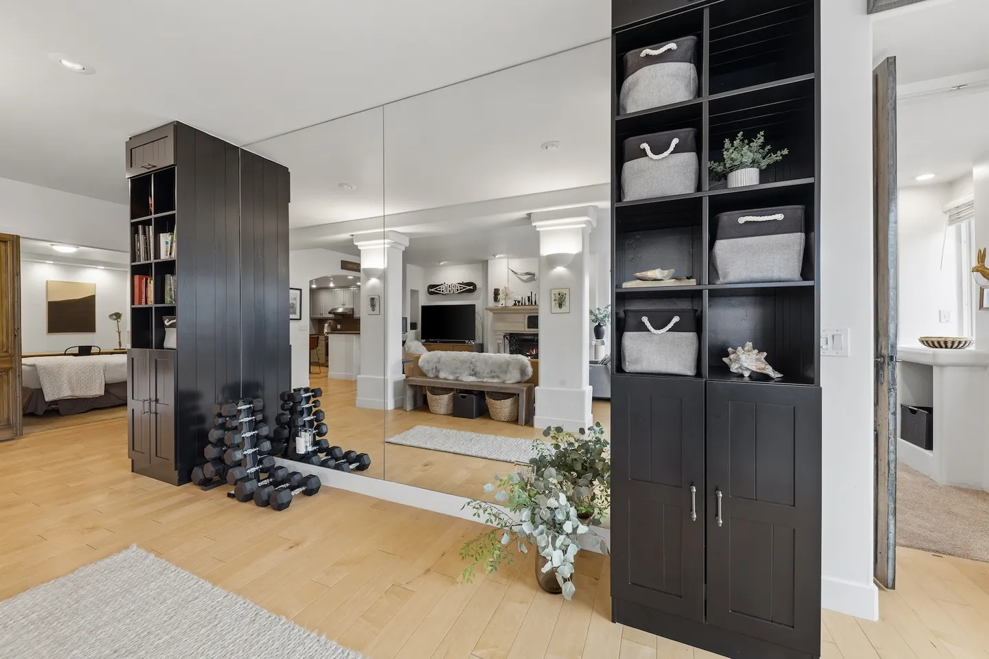 A home workout area featuring a large wall mirror, dark shelving unit with baskets, and a rack of dumbbells.