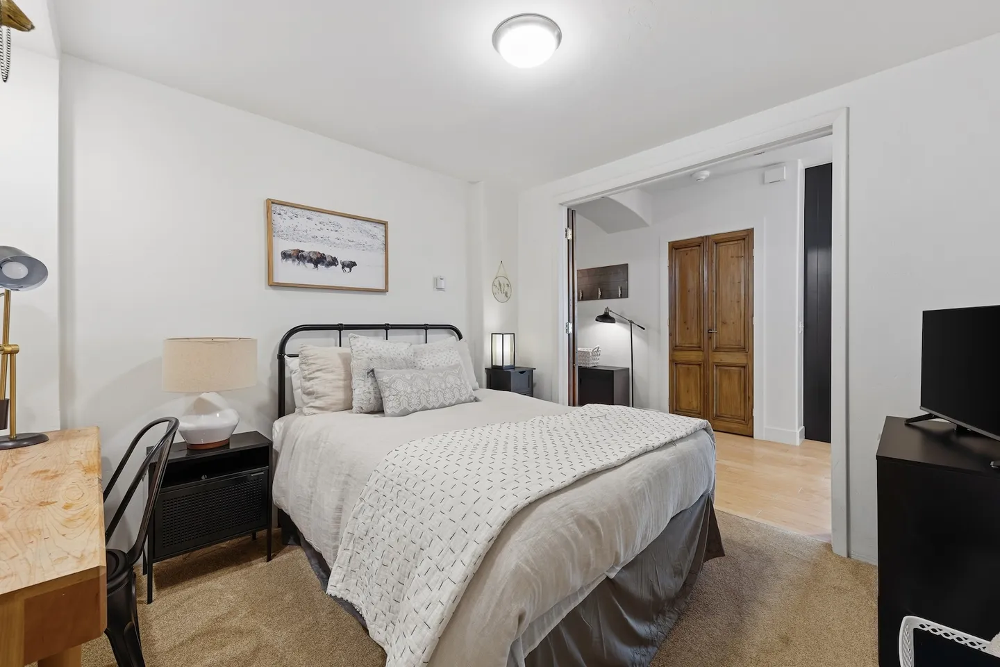 Bedroom with a metal-frame bed, bedside tables with lamps, a small desk, a television, and an open doorway leading to a hallway.