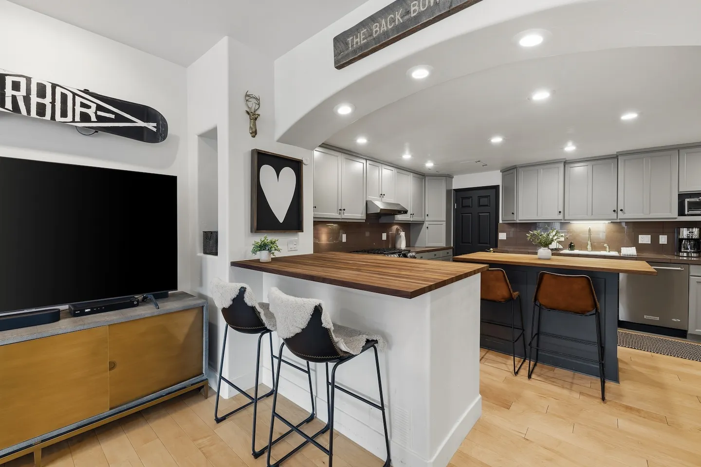 Open-concept living and kitchen area featuring a breakfast bar with two stools, a TV mounted on a media console, and an arched entry to the kitchen.