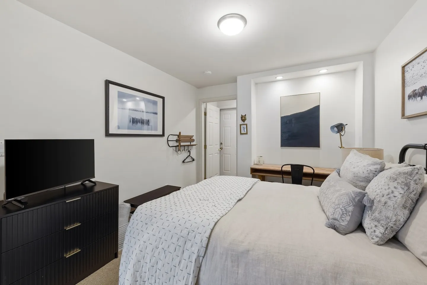 Bedroom featuring a made bed with pillows, a black dresser with a television, and a desk nook with recessed lighting.