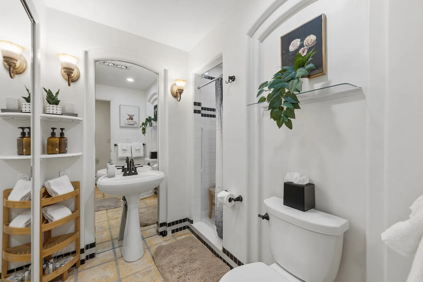 Bathroom with a white pedestal sink, arched wall mirror, toilet, tiled floor, and walk-in shower.