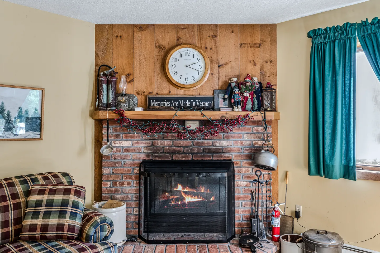 A corner brick fireplace with an active fire, wooden mantel, and wood paneling above with a wall clock.
