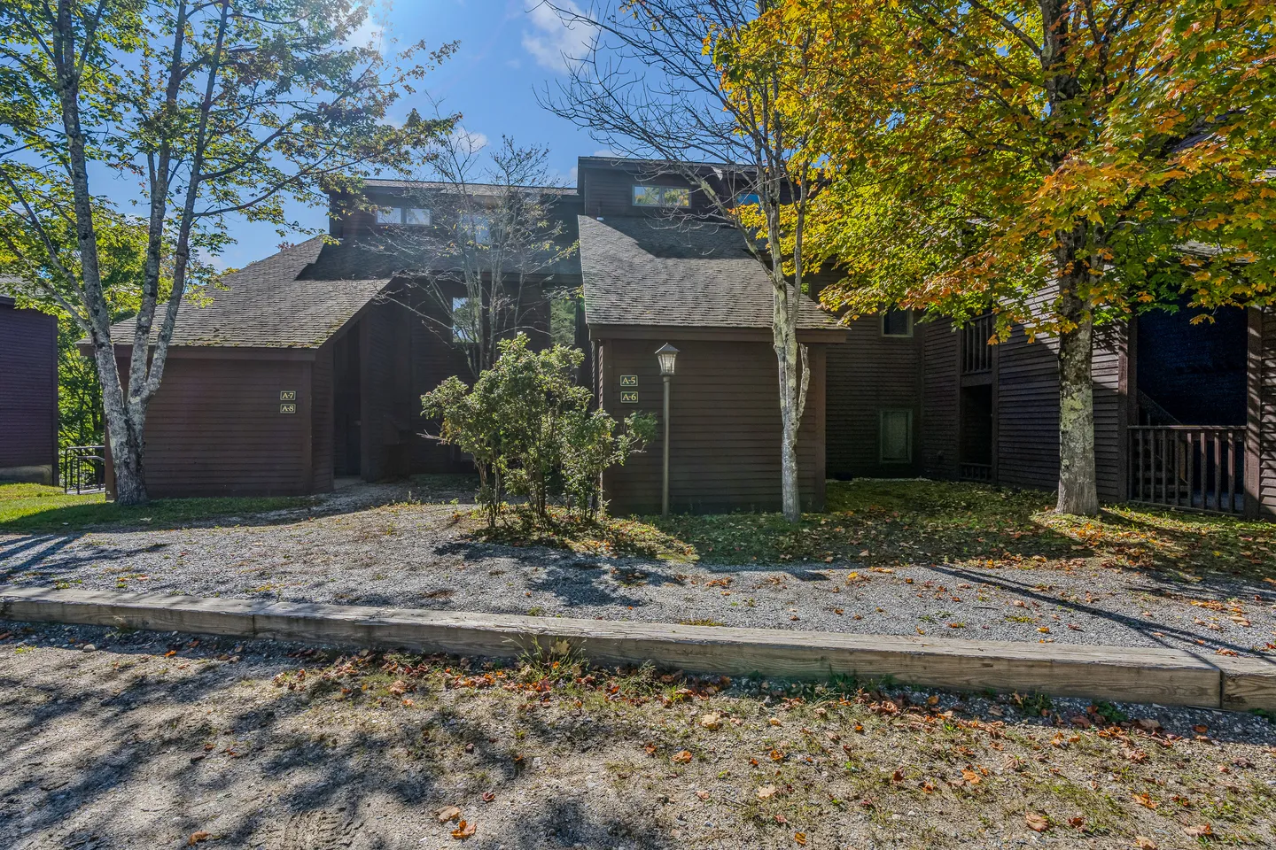 Exterior view of multi-level brown wood-sided townhome buildings with trees and gravel parking area.