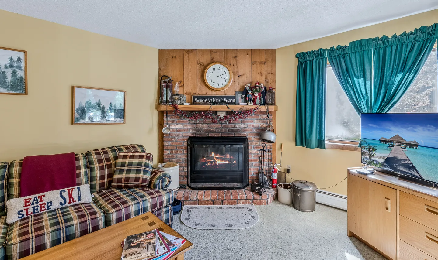 Living room with a plaid sofa, brick fireplace with lit fire, and a television set on a wood media stand.