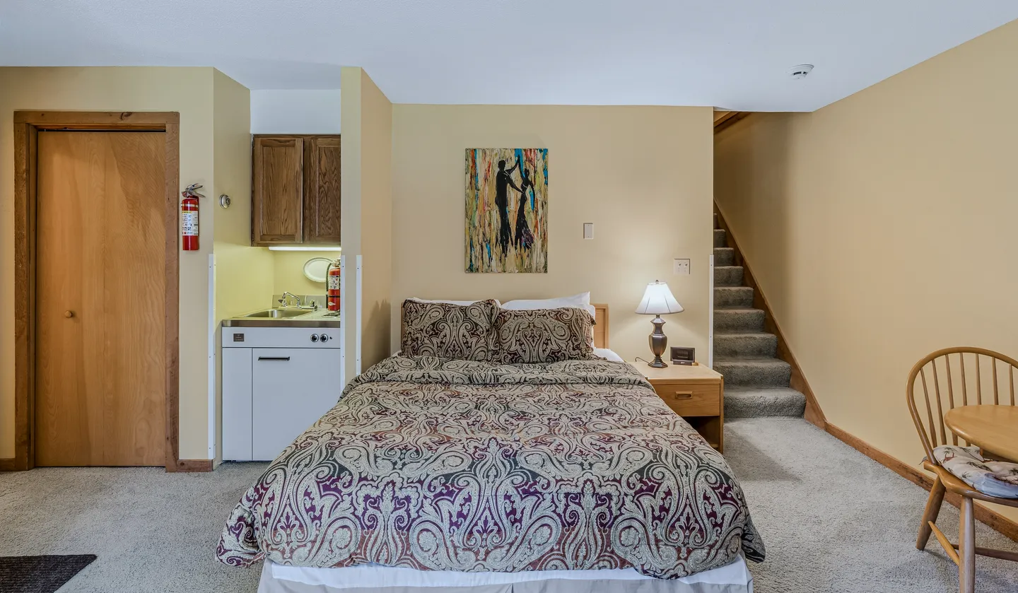Studio bedroom with a patterned bed, a compact kitchenette alcove to the left, and carpeted stairs to the right.