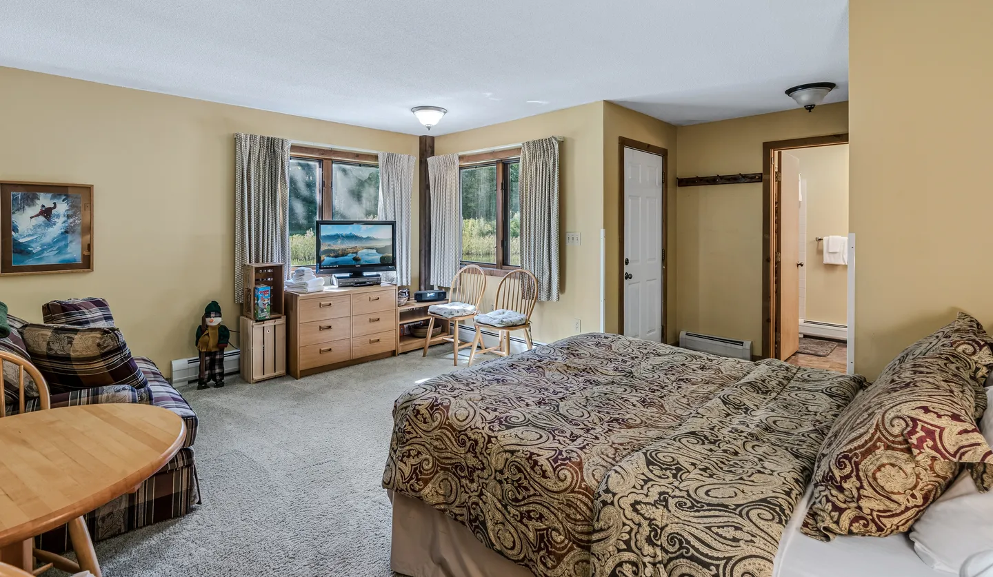 Bedroom with patterned bedding, carpeted floor, TV on a dresser, seating area, and an open doorway showing an attached bathroom.