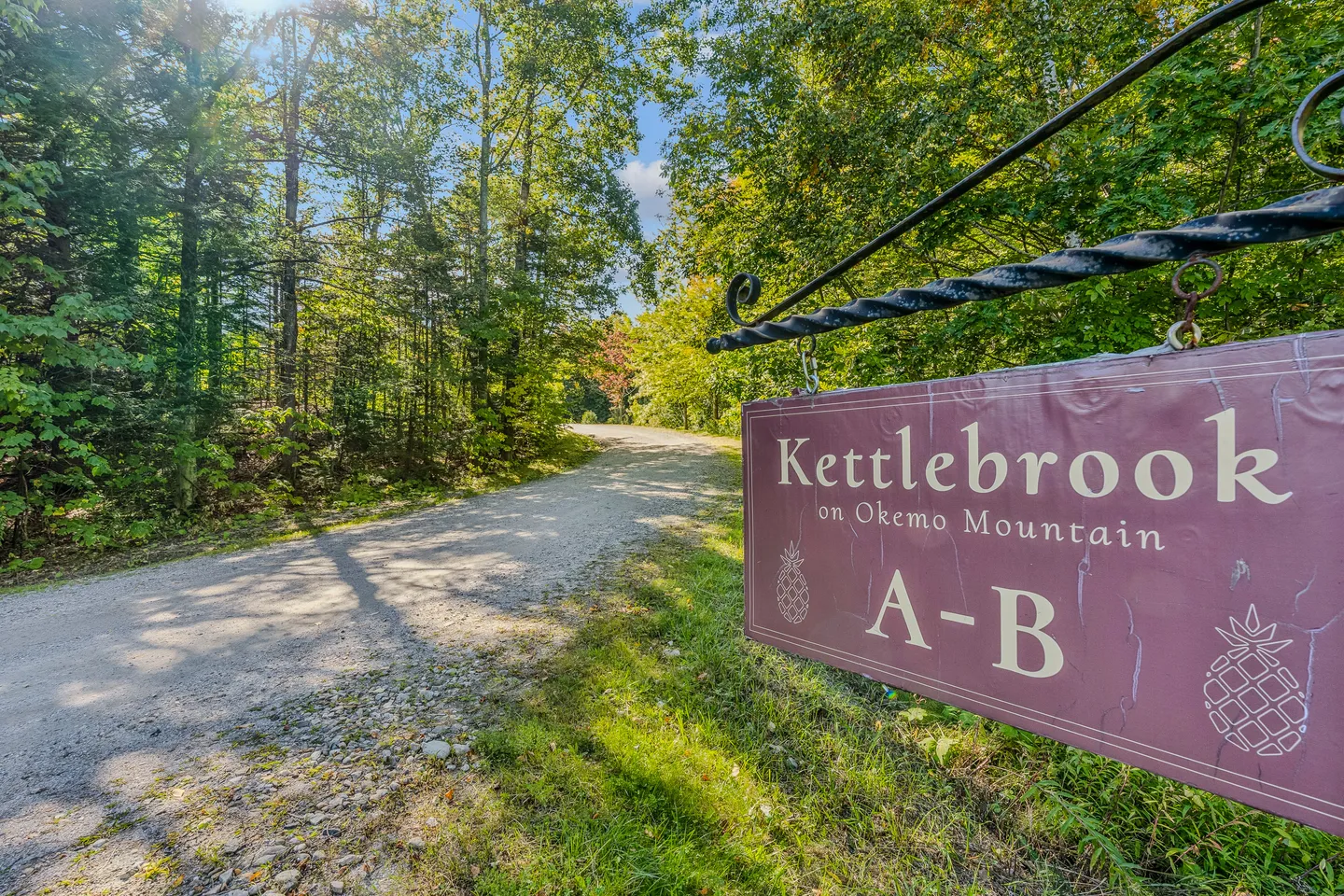 A maroon hanging sign reading Kettlebrook on Okemo Mountain A-B next to a tree-lined gravel road on a sunny day.