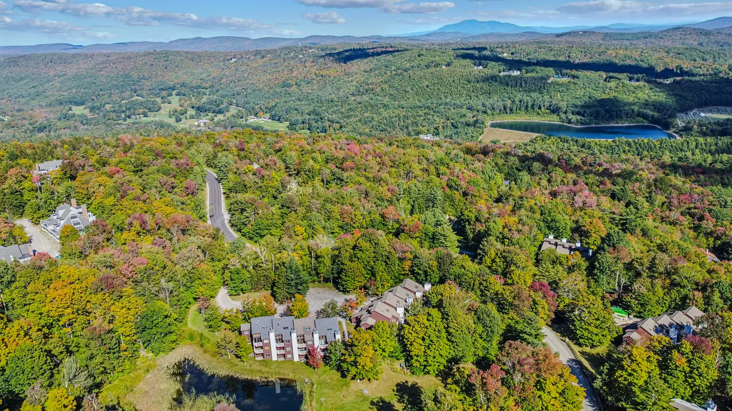 Aerial view of a mountain community surrounded by dense autumn foliage with distant hills, a lake, and a clear blue sky.