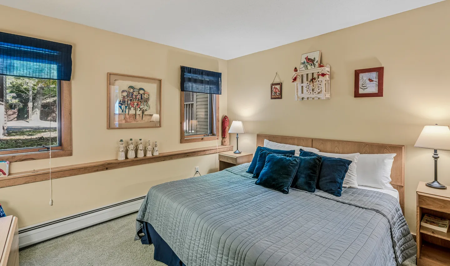 Bedroom with a wooden headboard bed made with a grey quilt, blue accent pillows, bedside tables with lamps, and carpeted flooring.