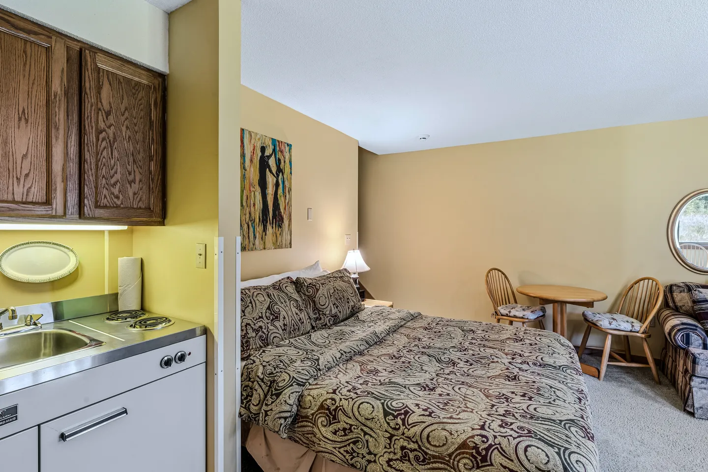 Studio interior showing a bed with patterned bedding next to a kitchenette counter, with a small dining table and chairs in the background.