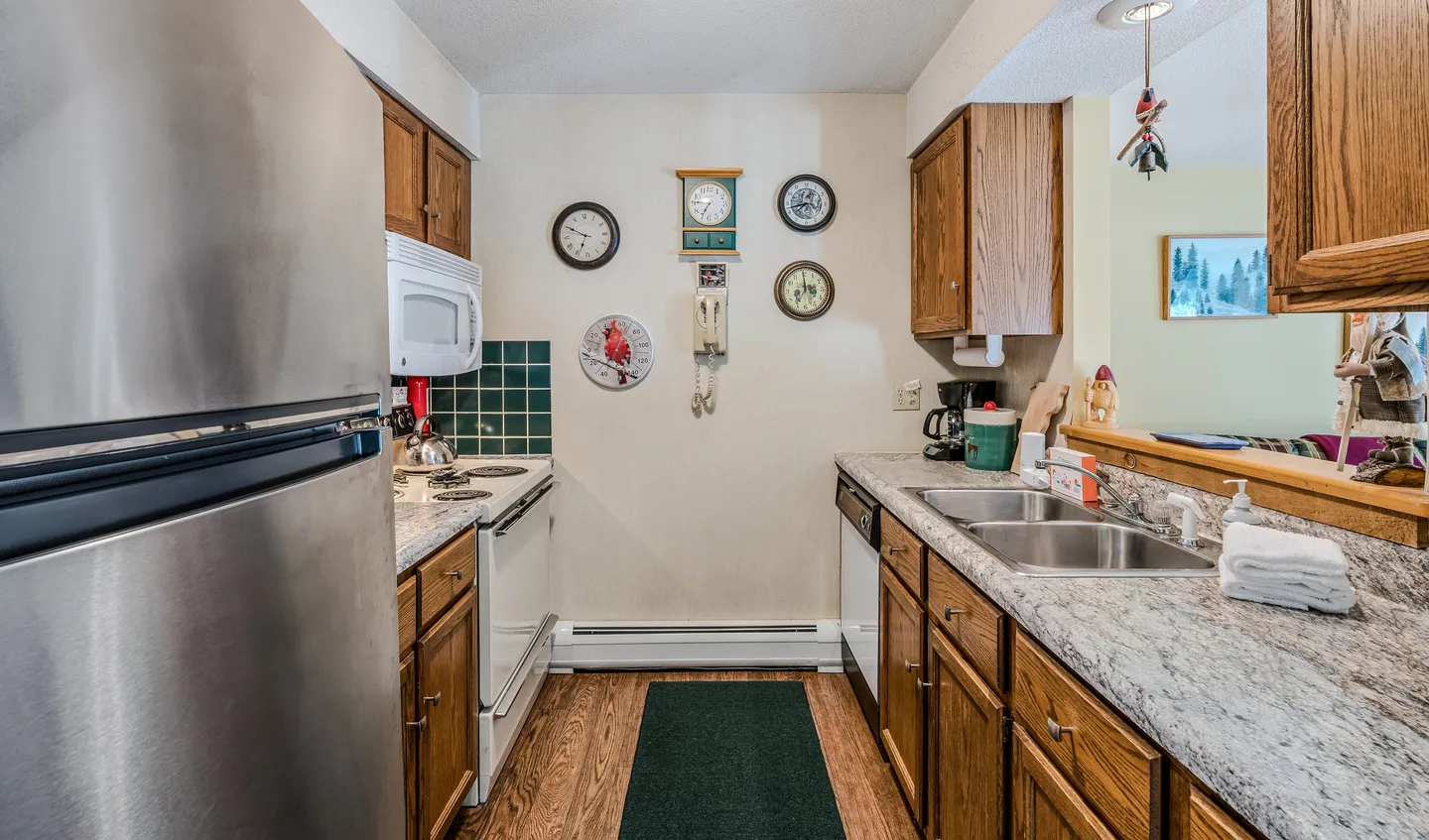 Galley-style kitchen with wood cabinets, light stone-patterned countertops, stainless steel refrigerator, and clocks on the back wall.
