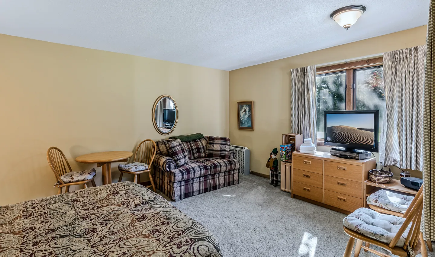 Bedroom with patterned bedding, a plaid loveseat, a small dining table with chairs, carpeted flooring, and a TV atop a wooden dresser.