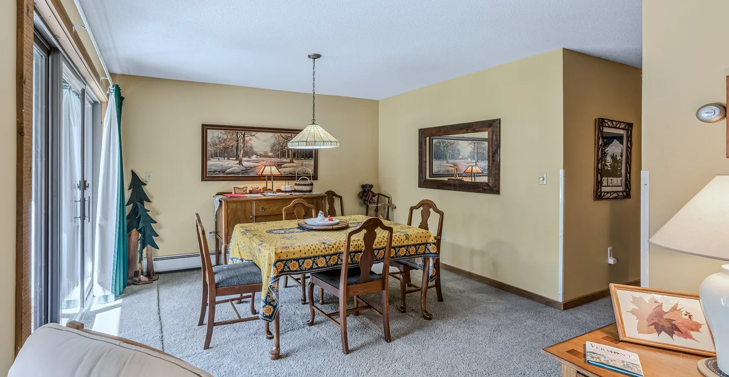 Dining area with a table covered by a patterned tablecloth, wooden chairs, carpet flooring, and a sliding glass door.