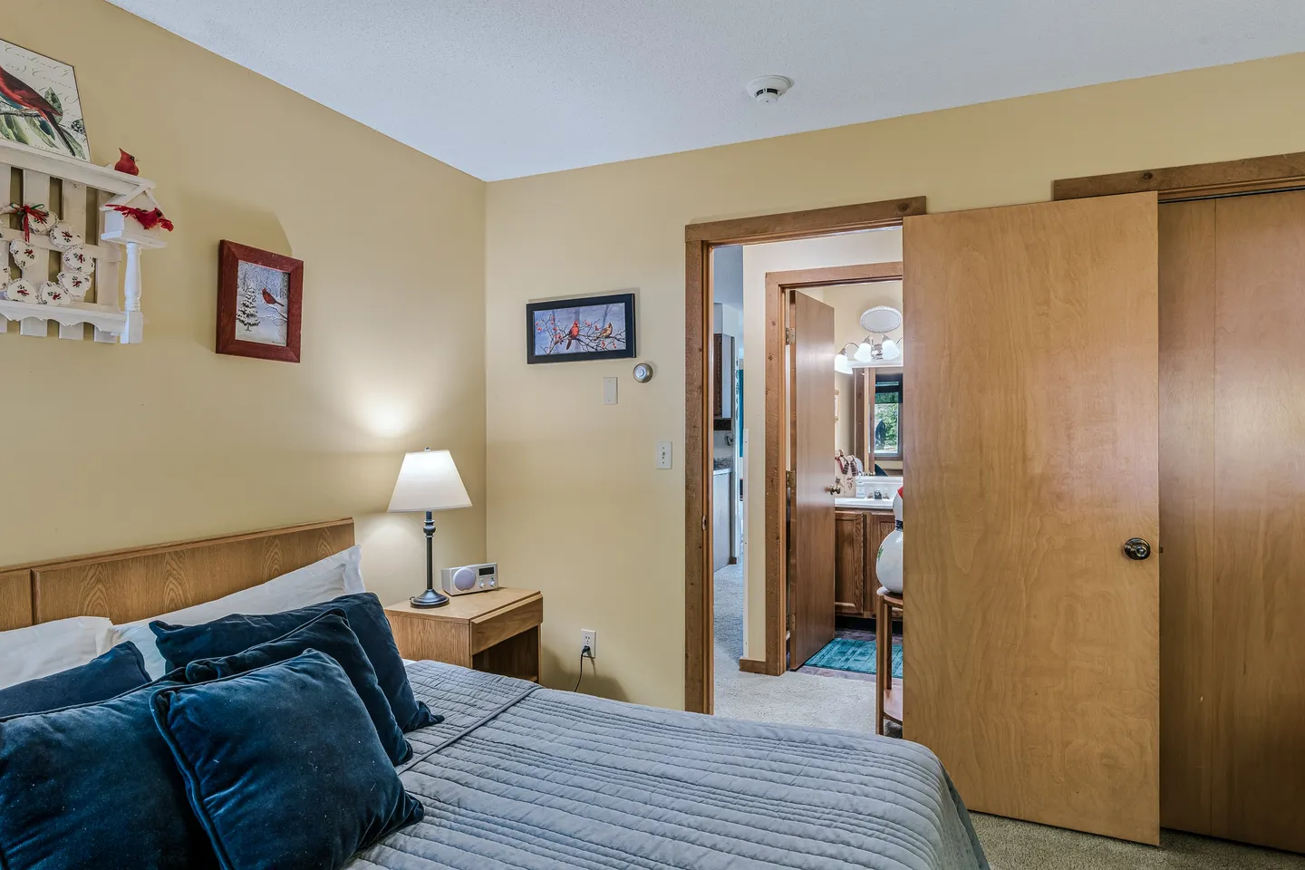 Bedroom featuring a made bed with navy blue pillows, a nightstand with a lamp, and an open doorway leading to an en-suite bathroom.