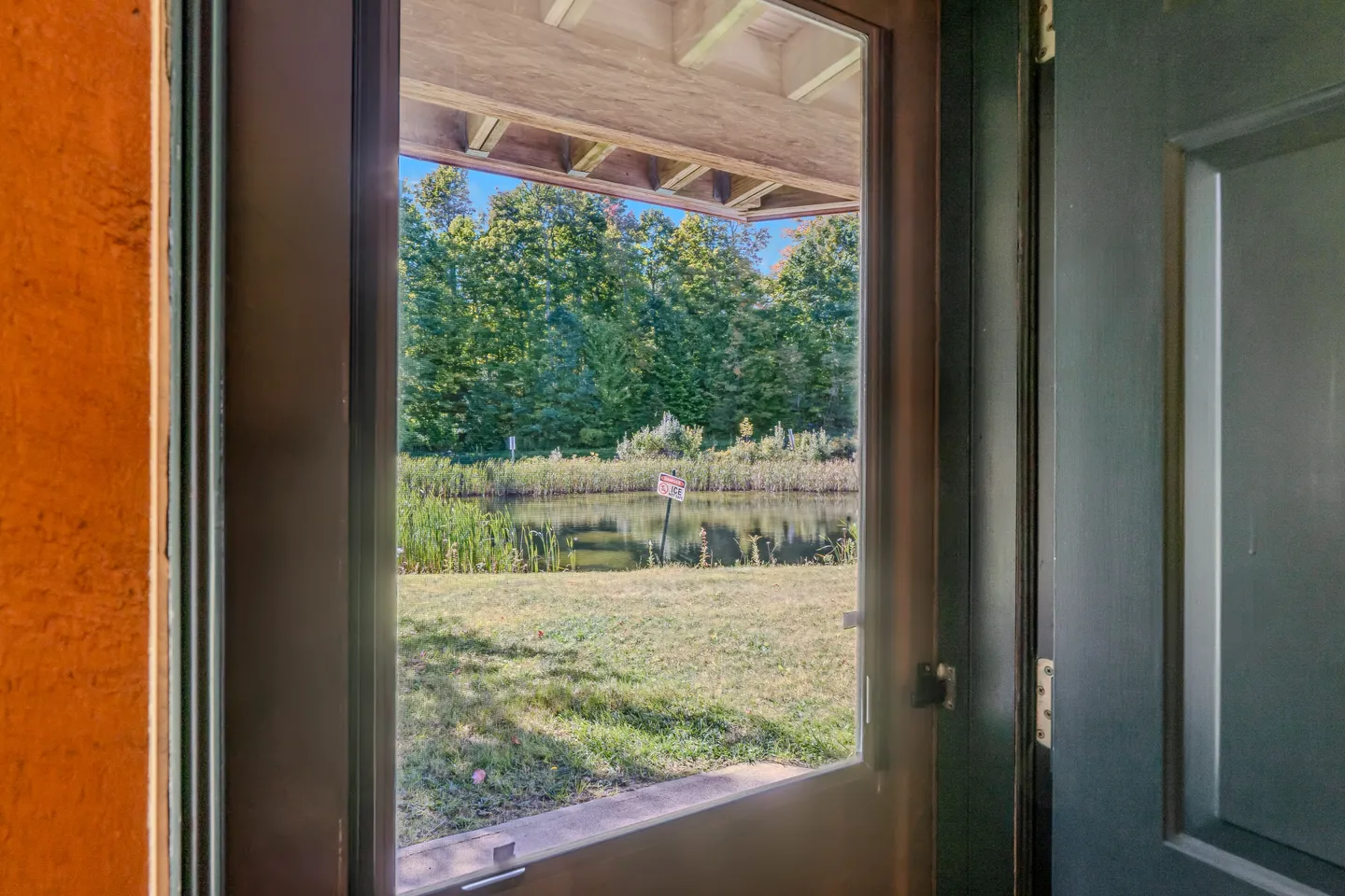 View through a glass storm door looking out at a grassy yard, small pond, and a dense line of green trees.