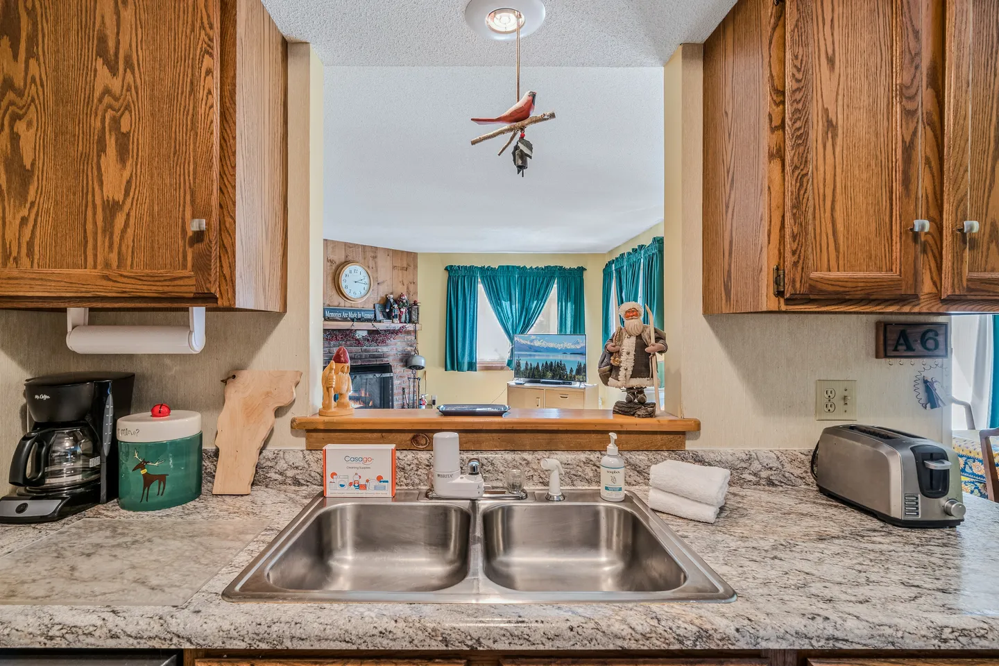 A kitchen sink area looking through a pass-through cutout into a living room with a fireplace and teal curtains.