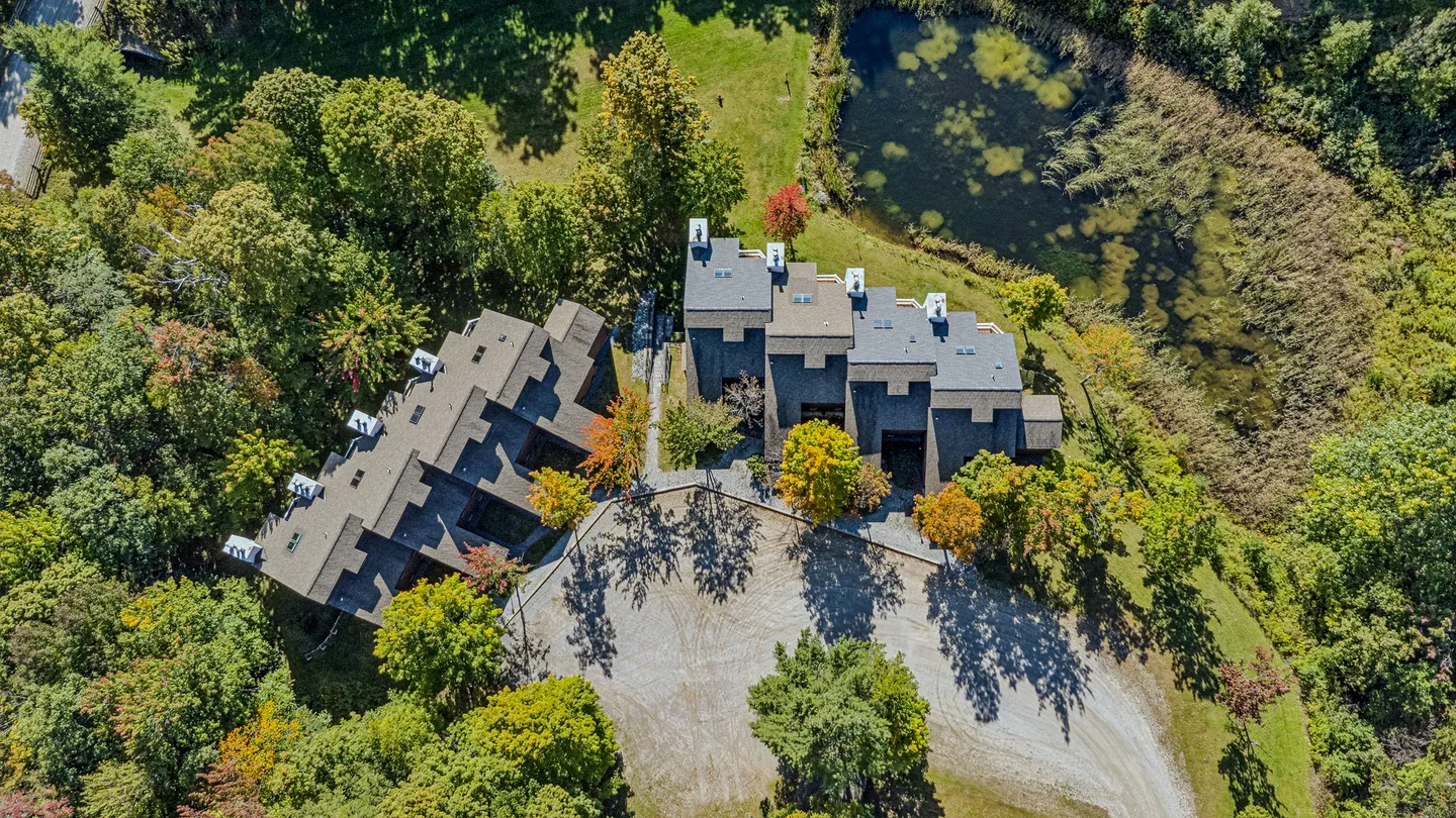 Aerial overhead view of multi-unit residential buildings surrounded by autumn trees, a gravel driveway, and a pond.