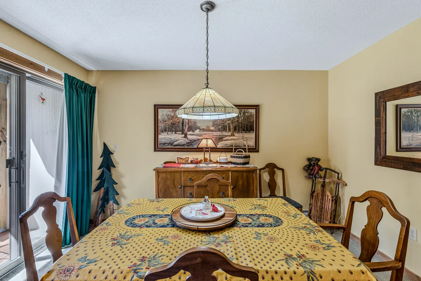 Dining area featuring a table covered with a patterned yellow tablecloth, wooden chairs, a hanging stained-glass pendant light, and a wooden buffet against a yellow wall.