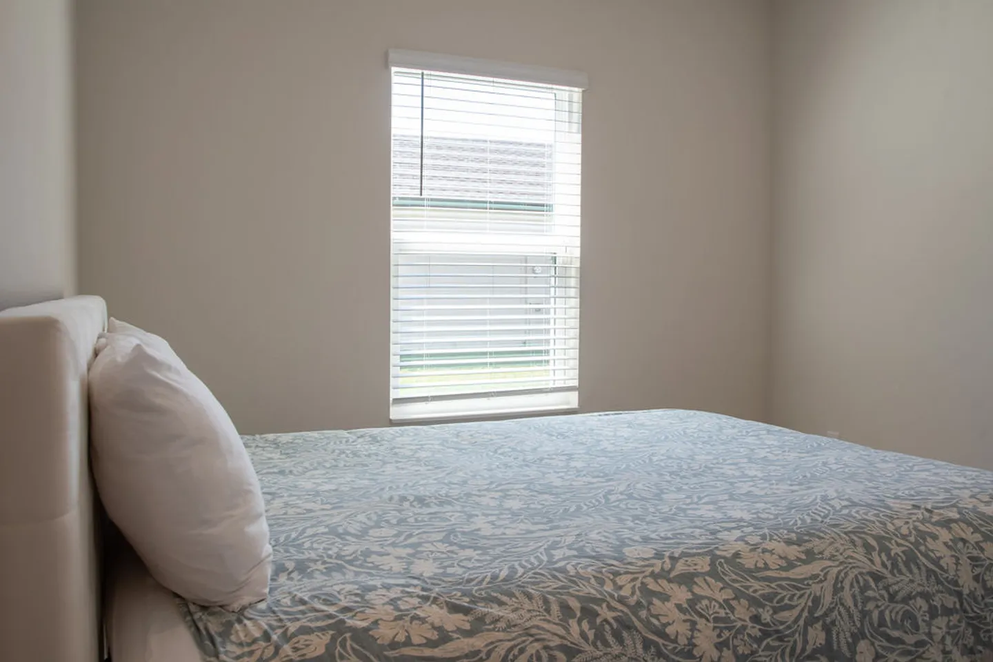 Bedroom corner showing a bed with a blue and white patterned bedspread beneath a window with horizontal blinds.