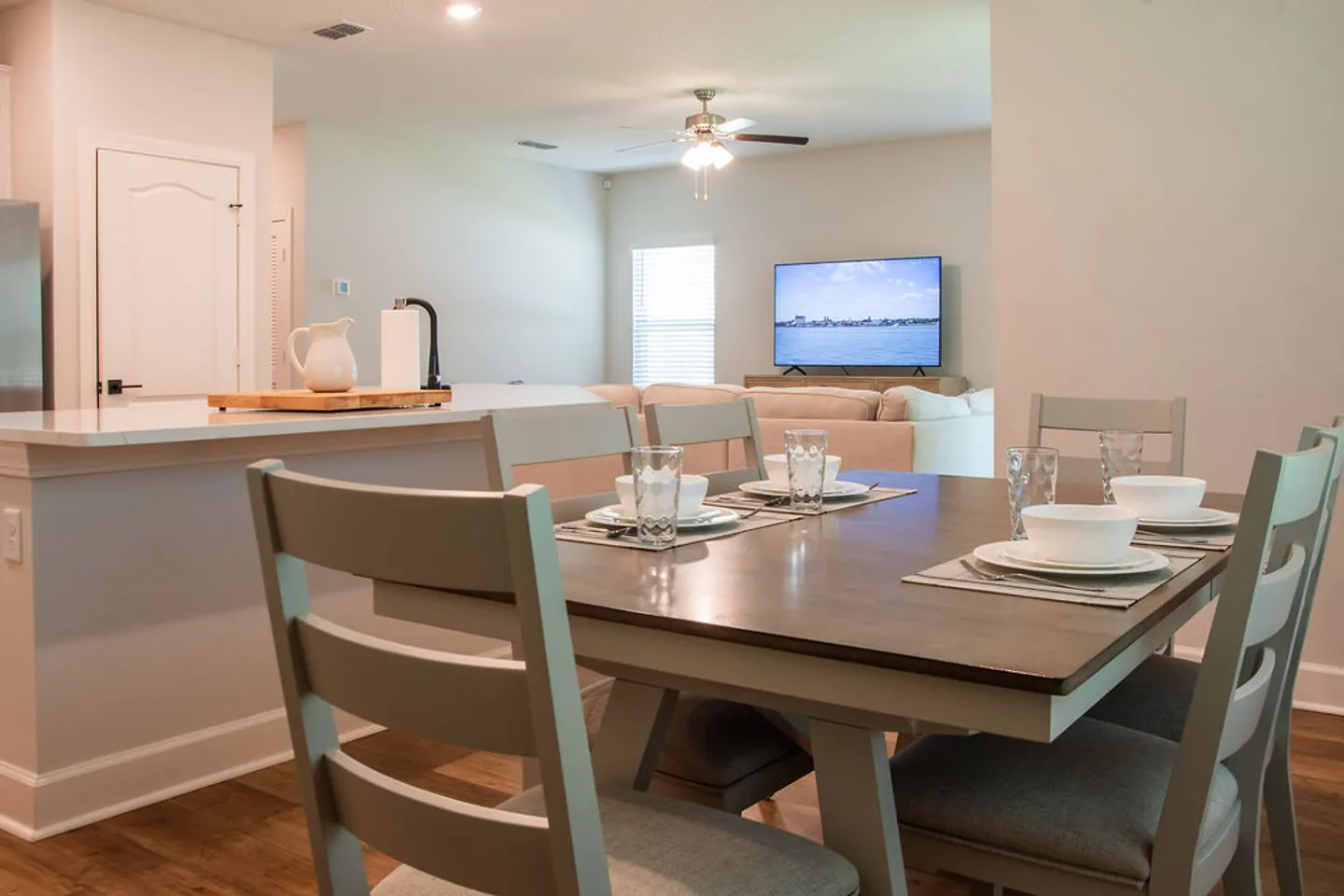 Dining area with a wooden table and set place settings, adjacent to an open-concept kitchen counter and living room with a television.