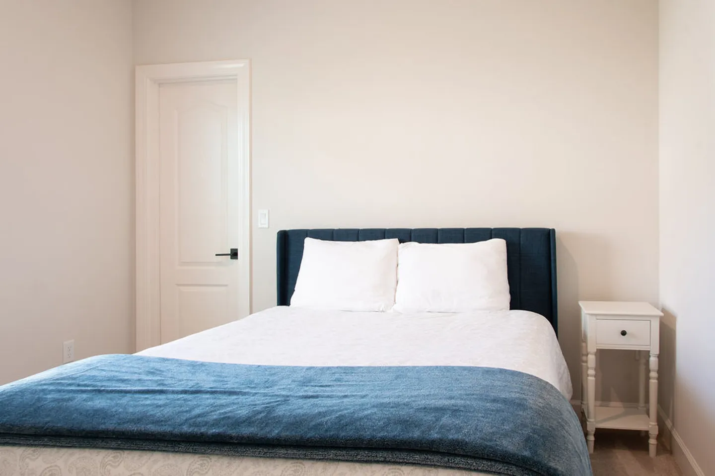Bedroom with a bed featuring a dark blue upholstered headboard, white bedding, a blue runner, and a white nightstand.