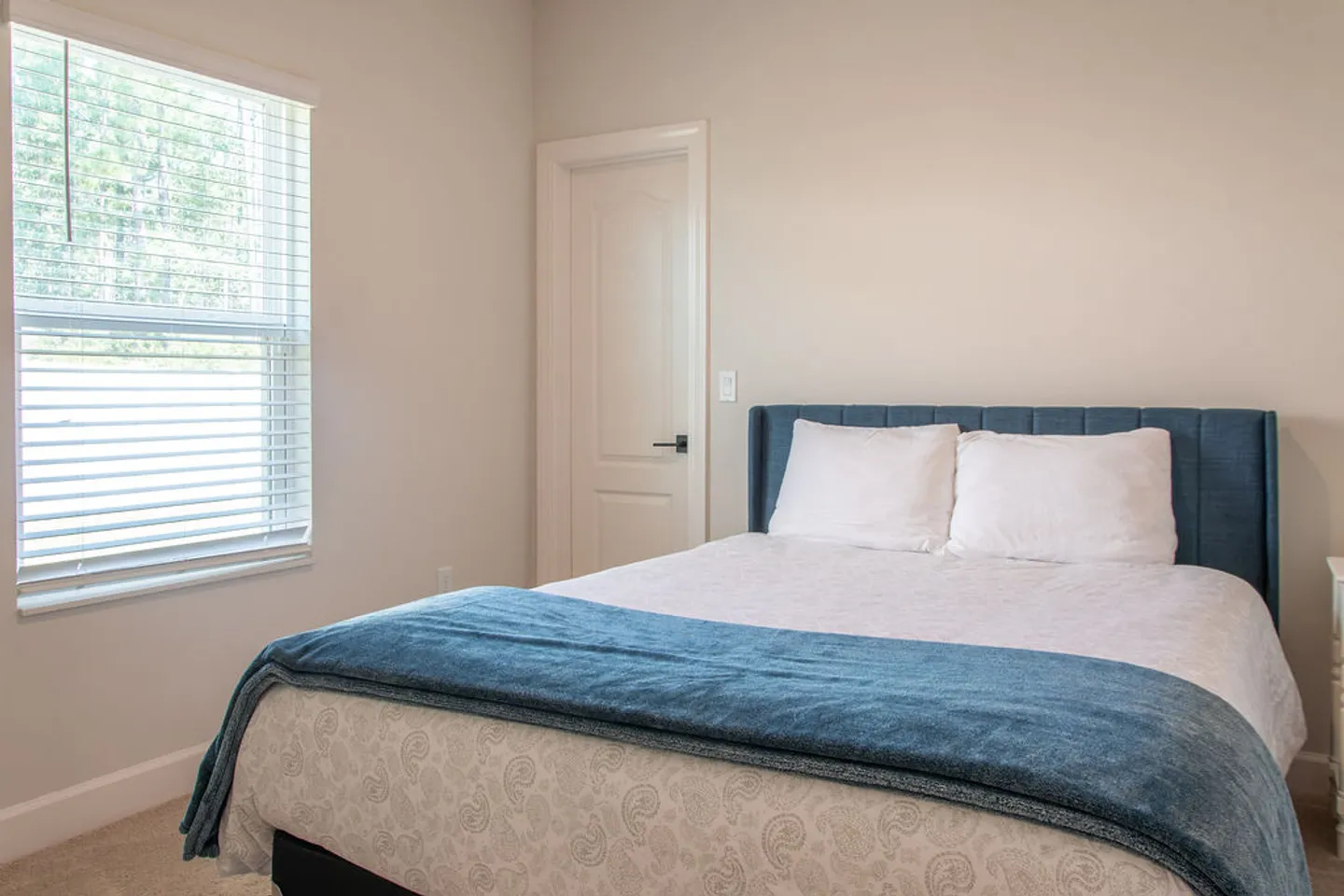 Bedroom featuring a made bed with an upholstered blue headboard and a window with white blinds.
