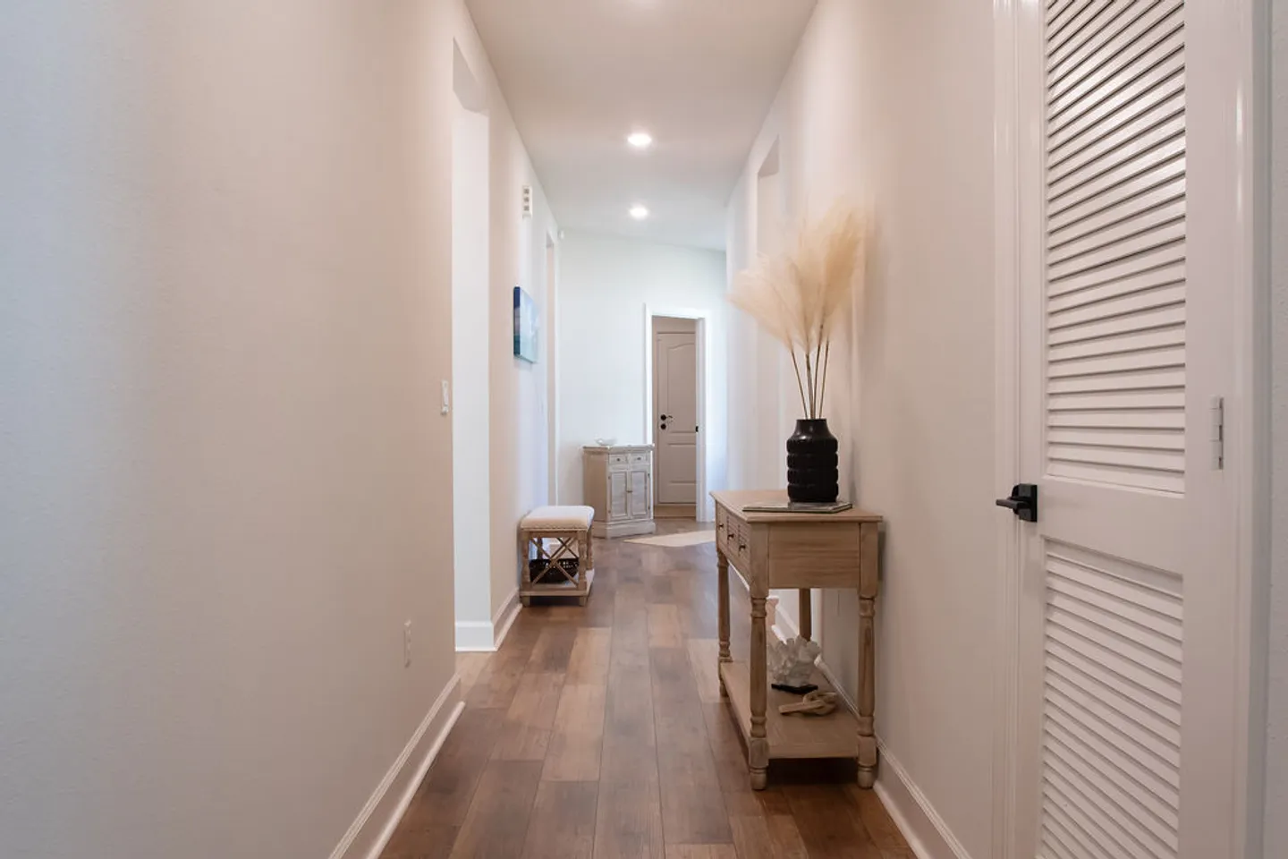 A long hallway with wood-look flooring, light-colored walls, recessed ceiling lights, and entryway furniture.