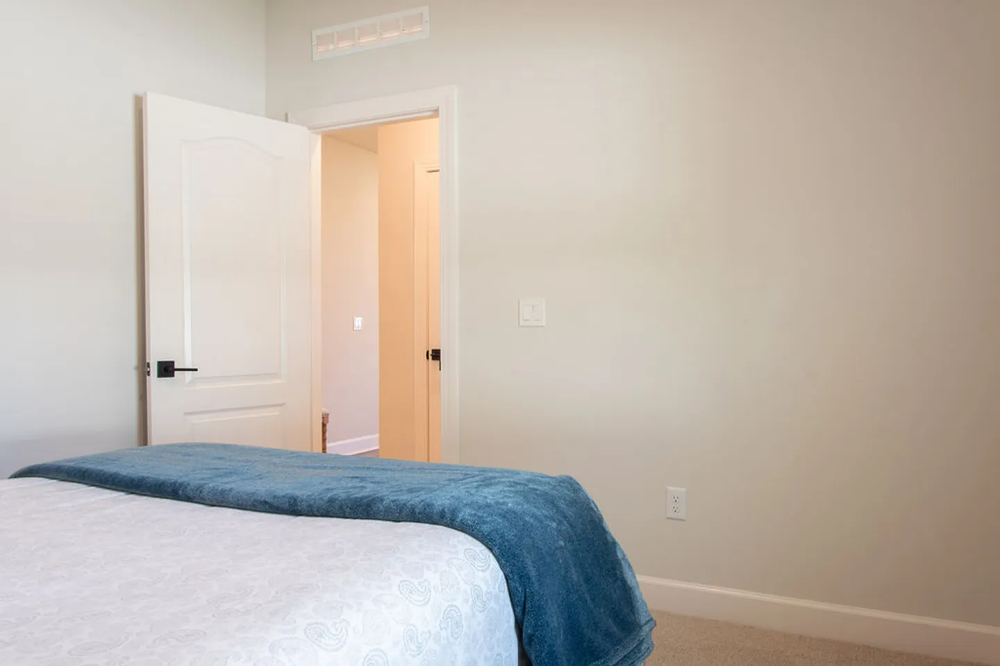 Bedroom corner showing the edge of a made bed with a blue throw blanket and an open interior door leading to a hallway.