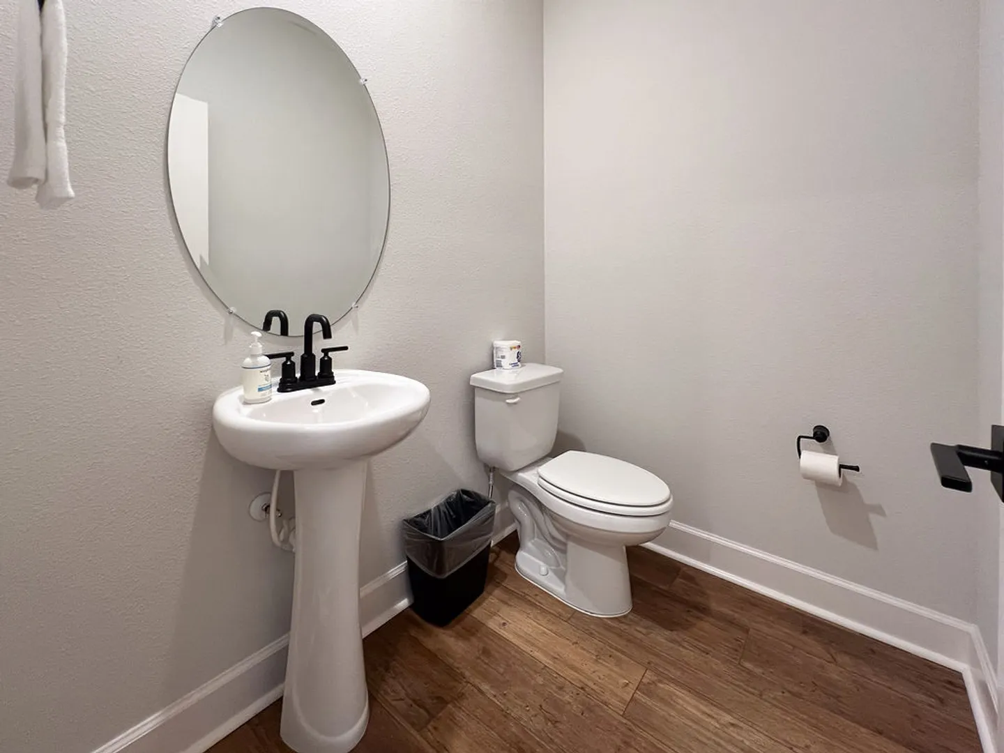 Powder room with a white pedestal sink, oval mirror, toilet, and wood-look flooring.