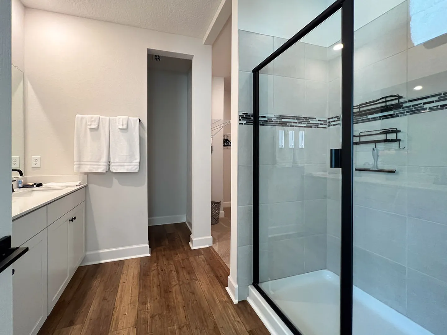 Bathroom showing a glass-enclosed walk-in shower with black framing, wood-look flooring, and a white vanity.